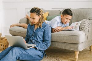 a woman looking at her laptop and a young boy reading a book on the couch behind her.