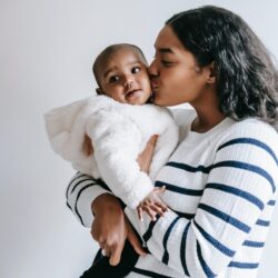 Black mother in striped shirt holding Black baby in fluffy white coat.