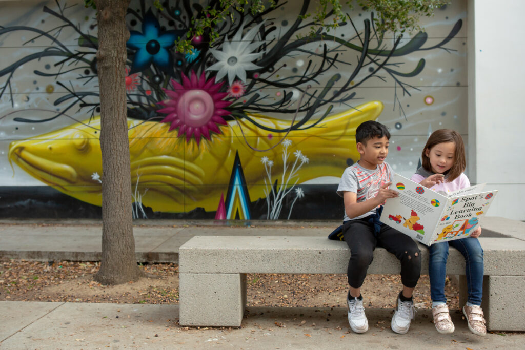 Two children sit on an outdoor bench reading a book.