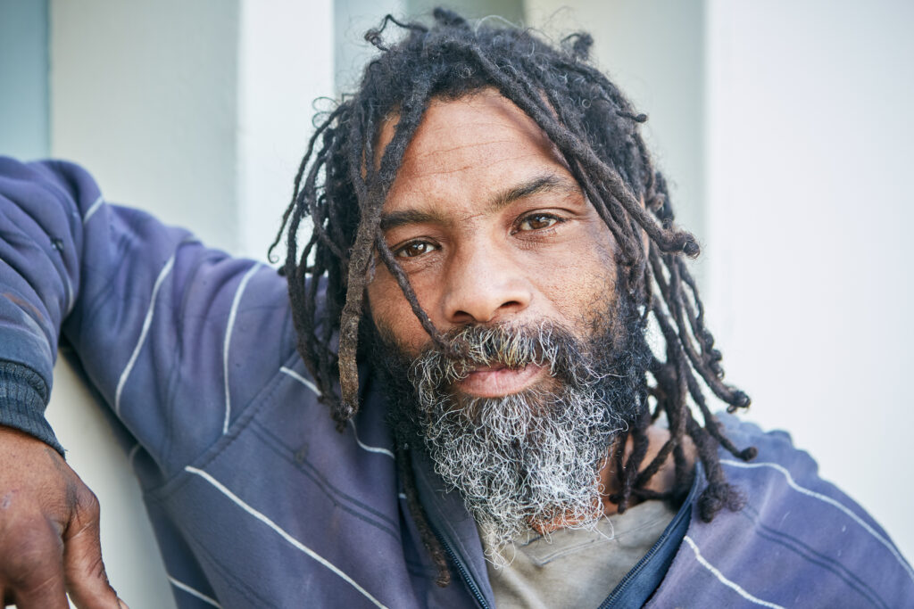 A homeless man with dreadlocks leans on a wall and looks at camera with a thoughtful but resigned expression.