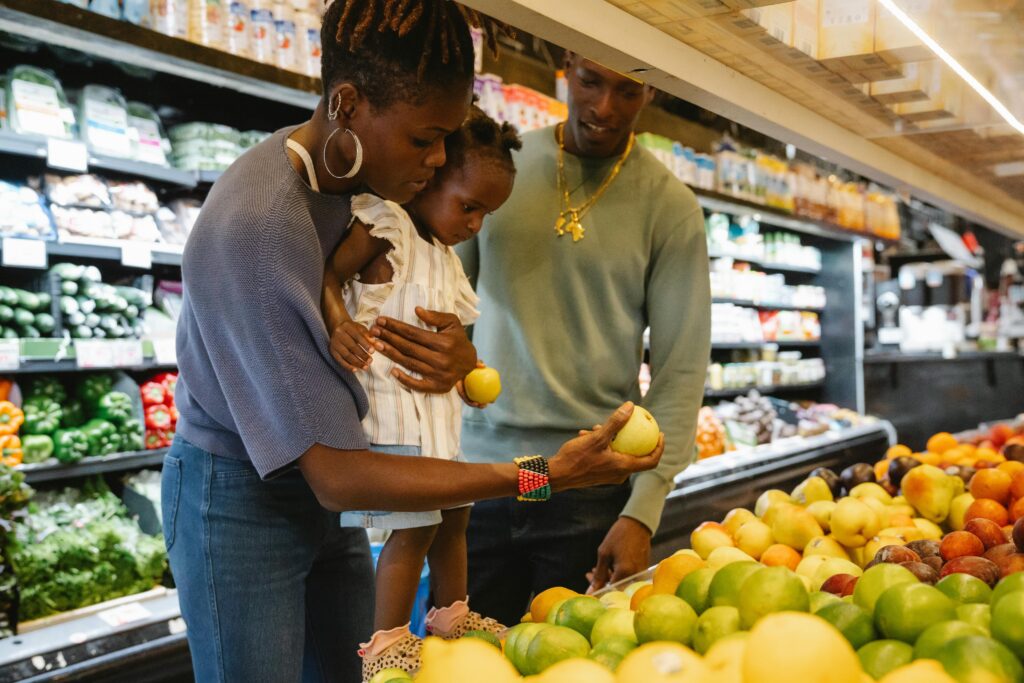 Photo of two Black parents and their child selecting produce at a grocery store