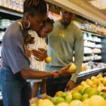 Photo of two Black parents and their child selecting produce at a grocery store