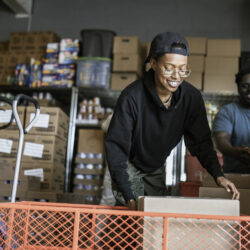 A young Black person lifts a box with shelves of other cardboard boxes behind them.