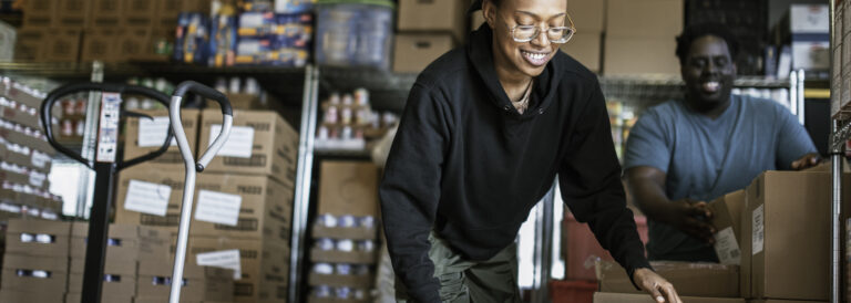 A young Black person lifts a box with shelves of other cardboard boxes behind them.